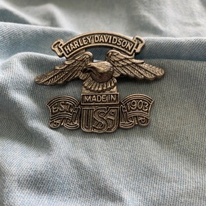 May include: A silver Harley Davidson pin featuring an eagle and the words "Harley Davidson" and "Made in USA" with the year "1903" at the bottom.