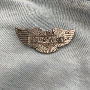 May include: A silver Harley-Davidson motorcycle wing pin with the words "Motor" and "Cycles" on the shield.