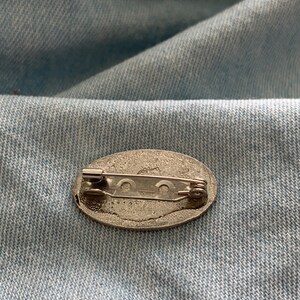 Puede incluir: Un broche de plata de forma ovalada con un cierre de seguridad en la parte posterior.