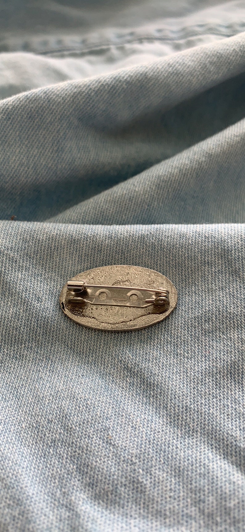 Puede incluir: Un broche de plata de forma ovalada con un cierre de seguridad en la parte posterior.