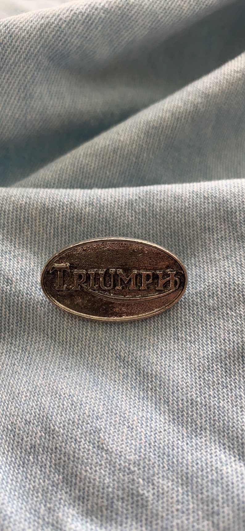 Puede incluir: Un pin de plata de forma ovalada con la palabra "TRIUMPH" en relieve. El pin est&aacute; sobre un fondo de mezclilla azul claro.