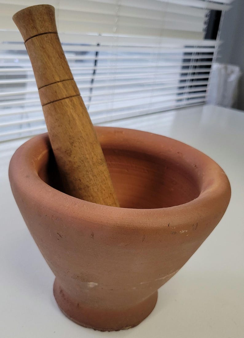 May include: A terracotta mortar and pestle set. The mortar is a wide-mouthed bowl with a rounded base. The pestle is made of wood and has a cylindrical shape with three carved rings near the top. The set is designed for grinding and mixing ingredients.