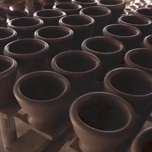 May include: A collection of brown ceramic mortar and pestle sets, arranged on wooden shelves. The mortars have a wide, open bowl shape, and the pestles are not visible. The lighting is soft, highlighting the texture of the pottery.