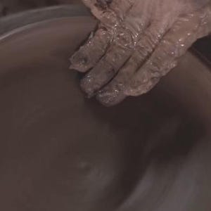 May include: Hands covered in wet clay shaping a pottery piece on a spinning wheel. The clay is a dark brown color, and the hands are actively molding the form. The image captures the process of pottery making.