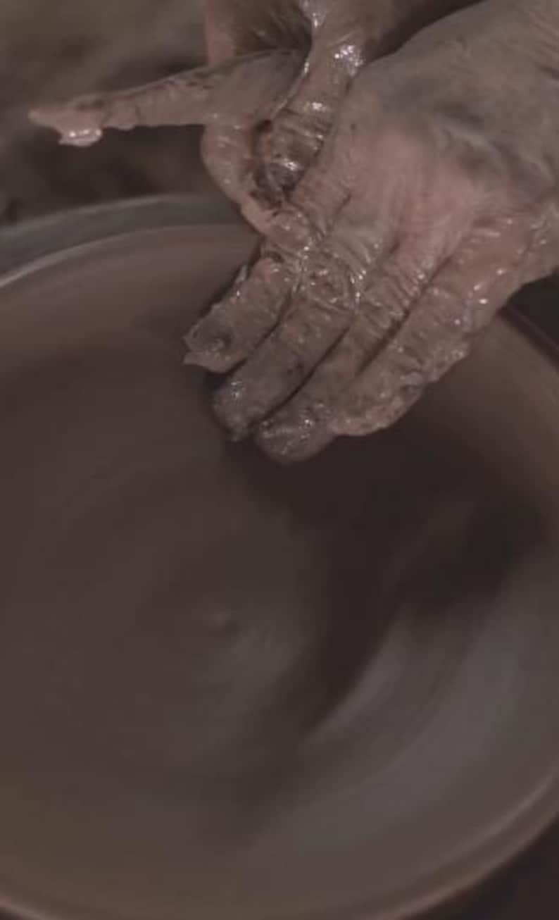 May include: Hands covered in wet clay shaping a pottery piece on a spinning wheel. The clay is a dark brown color, and the hands are actively molding the form. The image captures the process of pottery making.