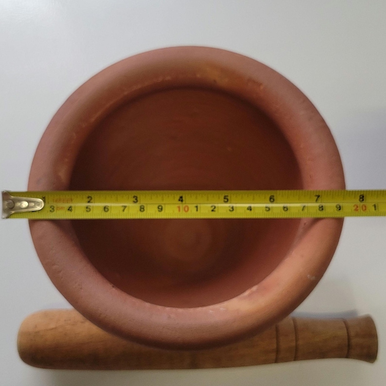 May include: A terracotta mortar and pestle set. The mortar is round, with a diameter of approximately 8 inches. A wooden pestle rests beside the mortar. The set is designed for grinding and mixing ingredients.