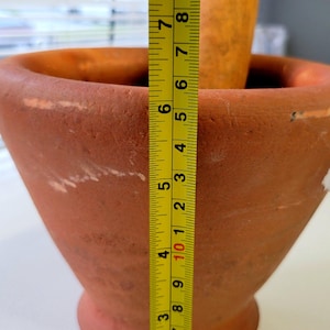 May include: A terracotta mortar and pestle set. The mortar is approximately 8 inches tall. The pestle is made of wood. The mortar has a wide opening and a tapered body. The set is designed for grinding and mixing ingredients.