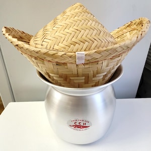 以下が含まれることがあります： 竹製の伝統的な炊飯器。編み込まれたバスケットの蓋と銀色の金属製の鍋のベースがあります。 蓋は円錐形で、より幅広のフレアバスケットの内側に収まります。 鍋にはテキスト入りの赤と白のステッカーが付いています。