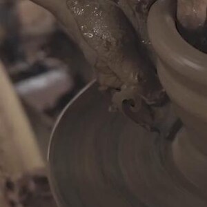 May include: A close-up shot of hands covered in wet clay shaping a pottery piece on a spinning wheel. The hands are molding the clay, creating a rounded form. The pottery wheel is in motion, and the clay is a light brown color.