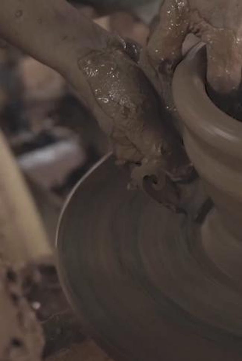 May include: A close-up shot of hands covered in wet clay shaping a pottery piece on a spinning wheel. The hands are molding the clay, creating a rounded form. The pottery wheel is in motion, and the clay is a light brown color.