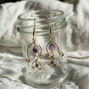 May include: A pair of gold wire earrings featuring amethyst and smaller purple beads. The earrings are dangling and have a delicate, whimsical design.
