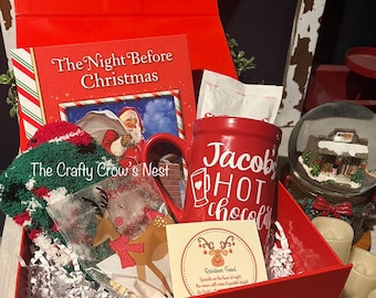 Może przedstawiać: Czerwone pudełko prezentowe wypełnione przedmiotami o tematyce świątecznej. Zawiera książkę "The Night Before Christmas", czerwony kubek z napisem "Jacob's Hot Chocolate", kartę z motywem renifera oraz zielono-czerwoną dzianinową czapkę.