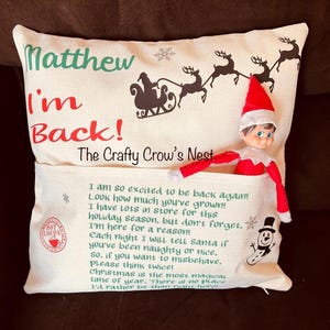 Puede incluir: Un cojín decorativo beige con un bolsillo, una pequeña muñeca de elfo y texto impreso que incluye "Matthew I'm Back!", "The Crafty Crow's Nest" y un poema navideño. El diseño incluye una silueta del trineo de Santa Claus y un muñeco de nieve.