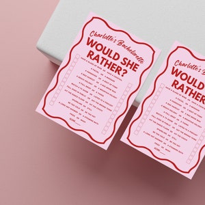 May include: Two pink cards with red text that says "Charlotte's Bachelorette Would She Rather?" with a list of questions to answer. The questions are in the format "Have a glass of wine or have a cocktail?" and "A facial or a massage?"