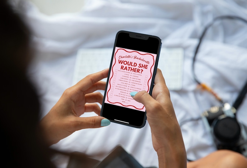 May include: A person is holding a smartphone displaying a pink and red "Would She Rather?" game for a hen party. The game is titled "Charlotte's Bachelorette Would She Rather?" and features a list of questions.