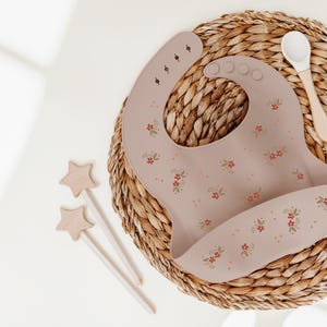 Peut inclure: Une bavette en silicone de couleur beige clair avec un motif floral et une petite poche. La bavette est posée sur une surface en osier tressé avec une cuillère en bois.