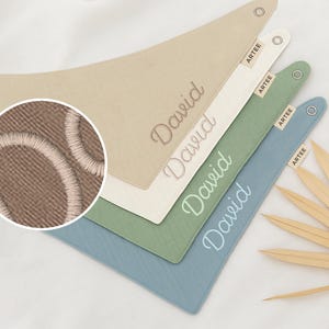 Può includere: Quattro bavaglini personalizzati per bambini in beige, bianco sporco, verde e blu. Ogni bavaglino è ricamato con il nome "David" in un colore coordinato. I bavaglini hanno una forma triangolare con chiusura a scatto e una piccola etichetta "ARTEE".