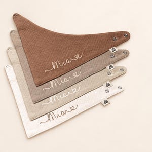 Könnte beinhalten: Ein Set aus vier dreieckigen Musselin-Lätzchen in den Farben Braun, Taupe, Beige und Creme. Jedes Lätzchen ist mit dem Namen Mia und einem kleinen Herz in Schreibschrift bestickt und hat ein kleines Stoffetikett mit einem Regenbogensymbol.