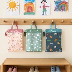 May include: Three personalised tote bags suspended from a wooden rail. The bags are pink, green, and teal, each bearing a name. The pink bag features a rainbow and star design, the green bag has a daisy pattern, and the teal bag displays an animal print.