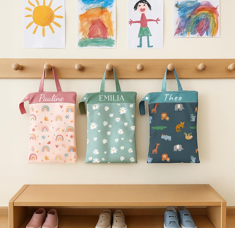 May include: Three personalised tote bags suspended from a wooden rail. The bags are pink, green, and teal, each bearing a name. The pink bag features a rainbow and star design, the green bag has a daisy pattern, and the teal bag displays an animal print.