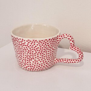 May include: White ceramic mug with a heart-shaped handle and a pattern of small red hearts. The mug has a wide opening and a smooth finish, ideal for tea or coffee.