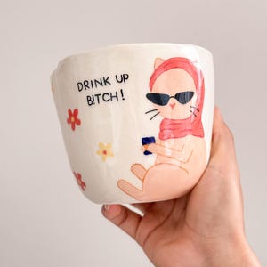 Pode incluir: Caneca de cerâmica branca com uma ilustração pintada à mão de um gato usando óculos de sol e um cachecol, segurando um pequeno objeto azul. A caneca tem o texto "DRINK UP BITCH!" e detalhes florais.