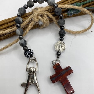 May include: A grey and black beaded keychain with a silver charm of the Virgin Mary and a red cross charm. The keychain has a silver clasp.