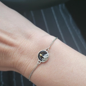 May include: Silver bracelet featuring a circular charm with a mountain scene and a yellow sun. The charm is set against a dark background. The bracelet is worn on a wrist, showcasing the delicate chain.