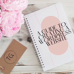 Könnte beinhalten: Ein weißes Spiralnotizbuch mit dem Titel "A Guide to: Creating a WordPress Website" in schwarzer Schrift und einem rosafarbenen Design. Ein braunes Etikett mit den Initialen "TG" und rosa Pfingstrosen sind ebenfalls zu sehen.