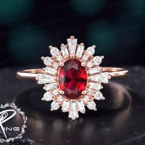Könnte beinhalten: Ein roségoldener Ring mit einem oval geschliffenen, tiefroten Edelstein im Zentrum, umgeben von einem Halo aus kleineren, klaren Edelsteinen und Baguette-Steinen. Der Ring hat ein elegantes Design und ein zartes Band.