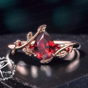 Könnte beinhalten: Ein Ring mit einem roten, birnenförmigen Edelstein, einem goldenen Band und filigranen Blatt- und Rankendetails. Der Ring ist mit kleinen roten Edelsteinen auf den Blättern verziert.