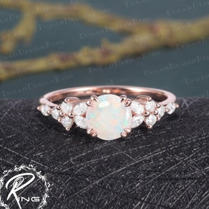 Könnte beinhalten: Ein Verlobungsring aus Roségold mit einem weißen Opal als Mittelstück und einem Cluster kleinerer Diamanten auf jeder Seite.