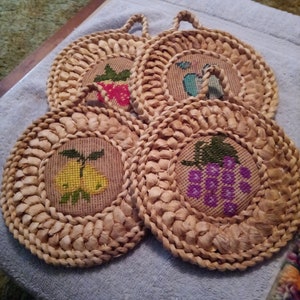 May include: Four round woven trivets with embroidered fruit designs: a strawberry, a pear, a bunch of grapes, and a fruit with a blue background. Each trivet has a woven handle. The trivets are made of natural-coloured woven material.