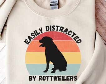 May include: A beige sweatshirt with a vintage-style graphic that says "Easily Distracted by Rottweilers". The graphic features a silhouette of a Rottweiler dog in front of a sunset.