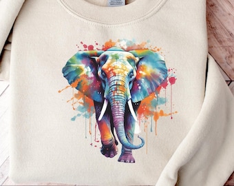 Puede incluir: Una sudadera beige con un gráfico de elefante acuarela colorido. El elefante está de frente con la trompa levantada. El gráfico tiene salpicaduras de rosa, azul, verde, naranja y amarillo.