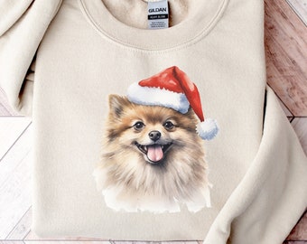 May include: A beige sweatshirt featuring a watercolor illustration of a Pomeranian dog wearing a Santa hat. The dog has brown and tan fur, a pink tongue, and is smiling. The sweatshirt has a crew neck and long sleeves.