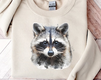 Peut inclure: Un sweat-shirt beige avec une illustration aquarelle d'un visage de raton laveur. Le raton laveur a les yeux noirs et le nez noir. Le sweat-shirt a une étiquette Gildan sur le devant.