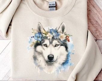 Może przedstawiać: Beżowa bluza z nadrukiem akwarelowej ilustracji psa husky w koronie kwiatów. Pies ma szare i białe futro, z niebieskimi i białymi kwiatami w koronie. Bluza jest złożona na drewnianej powierzchni.