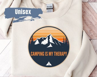 May include: A beige sweatshirt with a circular graphic of mountains and a sunset. The graphic has the text "Camping is my therapy" and a small tent icon.