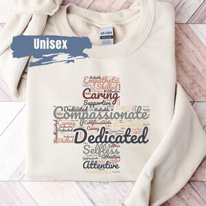 May include: A beige unisex sweatshirt with a heart-shaped word cloud design. The words in the cloud are: Empathetic, Skilled, Caring, Supportive, Dedicated, Reliable, Compassionate, Selfless, and Attentive.