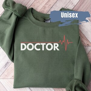 May include: A green unisex sweatshirt with the word "DOCTOR" printed in white with a red heartbeat graphic above it. The sweatshirt is folded and laid on a wooden surface.