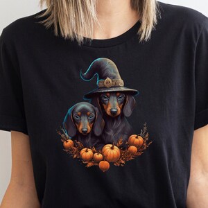 Może przedstawiać: Czarny t-shirt z ilustracją na Halloween. Grafika przedstawia dwa jamniki, z kt&oacute;rych jeden ma na sobie kapelusz czarownicy, otoczone dyniami i jesiennymi liśćmi. Projekt jest kolorowy i szczeg&oacute;łowy, idealny na straszny sezon.