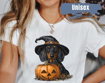 Op de afbeelding: Wit T-shirt met een zwart-bruine teckel met een heksenhoed, naast een oranje pompoen lantaarn. Het woord "Unisex" staat op het shirt. Een Halloween-thema ontwerp.