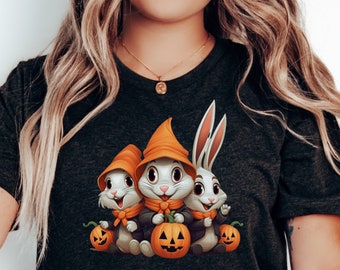 Könnte beinhalten: Dunkelgraues T-Shirt mit einem Halloween-Motiv. Die Grafik zeigt drei Cartoon-Hasen mit orangefarbenen Hexenhüten, die geschnitzte Kürbisse halten. Die Hasen tragen schwarze Jacken und orangefarbene Schals.
