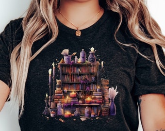 May include: Dark gray t-shirt featuring a detailed illustration of a mystical bookshelf. The design includes stacks of books, candles, potion bottles, and other magical elements in shades of purple, gold, and brown. The overall theme is fantasy and witchcraft.