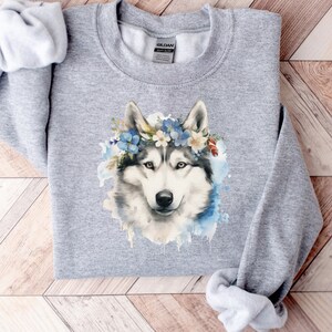 Op de afbeelding: Een lichtgrijs sweatshirt met een aquarel illustratie van een husky hond met een bloemenkrans. De vacht van de hond is wit en grijs, met blauwe en witte bloemen in de krans. De sweatshirt heeft een ronde hals en lange mouwen.
