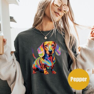 May include: A dark gray sweatshirt with a colorful dachshund illustration. The dachshund is painted in a rainbow of colors and has a brown nose. The text "Pepper" is printed in yellow on the bottom right corner of the image.