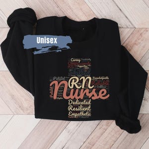 May include: Black sweatshirt with a word cloud design featuring the word "Nurse" in large, pink letters. The word cloud includes words like "caring", "compassionate", "skilled", "patient", "dedicated", "resilient", and "empathetic".