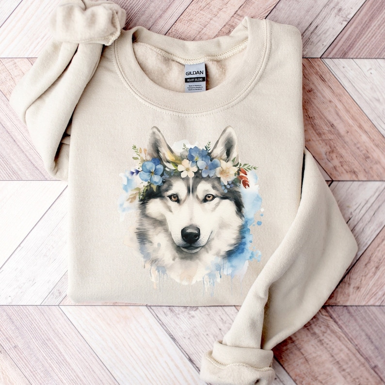 Op de afbeelding: Beige sweatshirt met een aquarel illustratie van een husky hond met een bloemenkrans. De hond heeft grijs en wit haar, met blauwe en witte bloemen in de krans. De sweatshirt is op een houten oppervlak gevouwen.