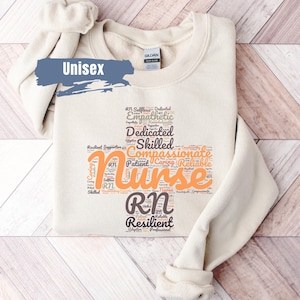 May include: A beige unisex sweatshirt with a word cloud design featuring the word "Nurse" in orange. The word cloud includes words like "RN", "Selfless", "Dedicated", "Empathetic", "Skilled", "Compassionate", "Patient", "Caring", and "Resilient".